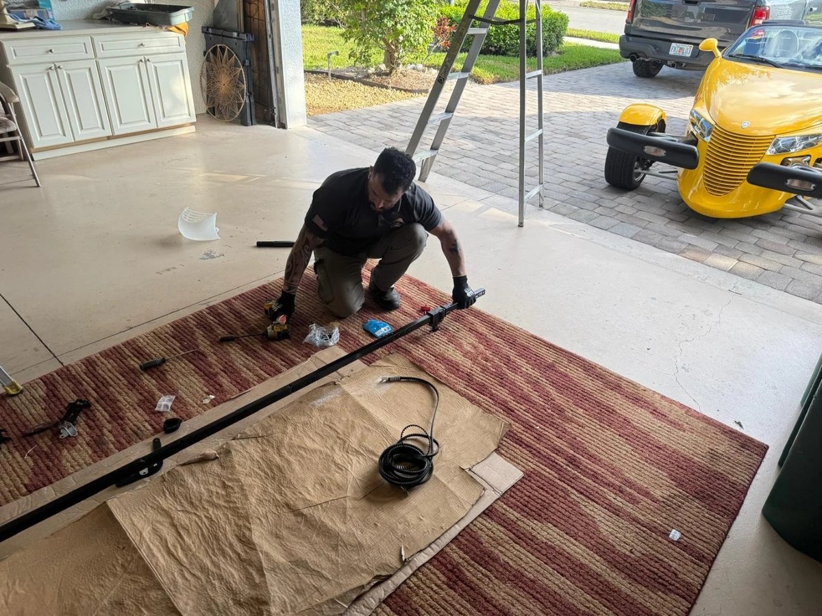 Technician kneeling on a rug beside a long steel shaft laid on the floor of a garage