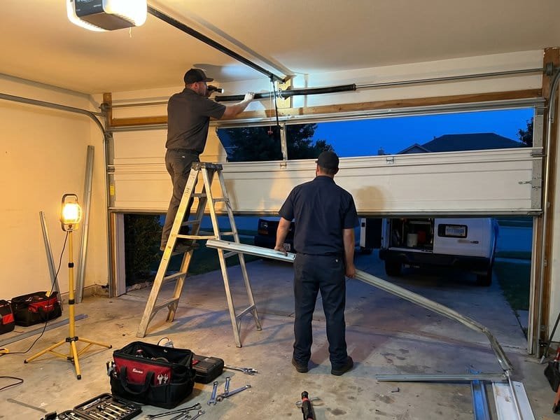 Two technicians working at dusk on a raised garage door lit by a portable work lamp